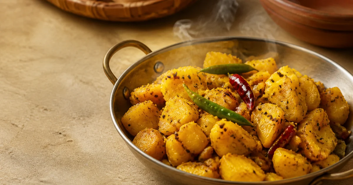 A shallow commercial tray of dry Bangladeshi alu bhaji with golden potato batons, sliced onions, green chillies and coriander.