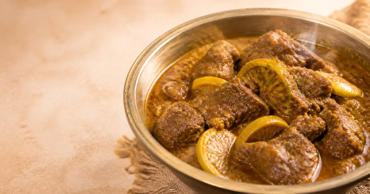 A serving bowl of Sylheti beef shatkora with tender beef pieces, thick spiced gravy and visible green citrus wedges.