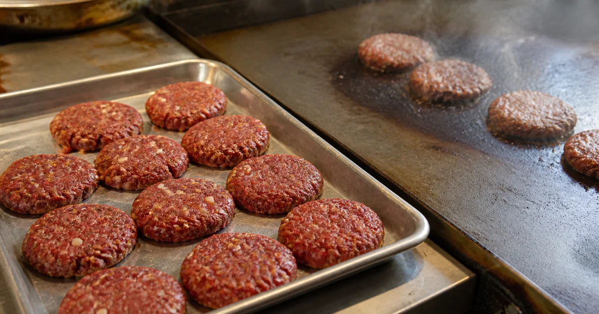 Forty evenly shaped beef burger patties separated with paper beside freshly griddled browned patties in a commercial kitchen.