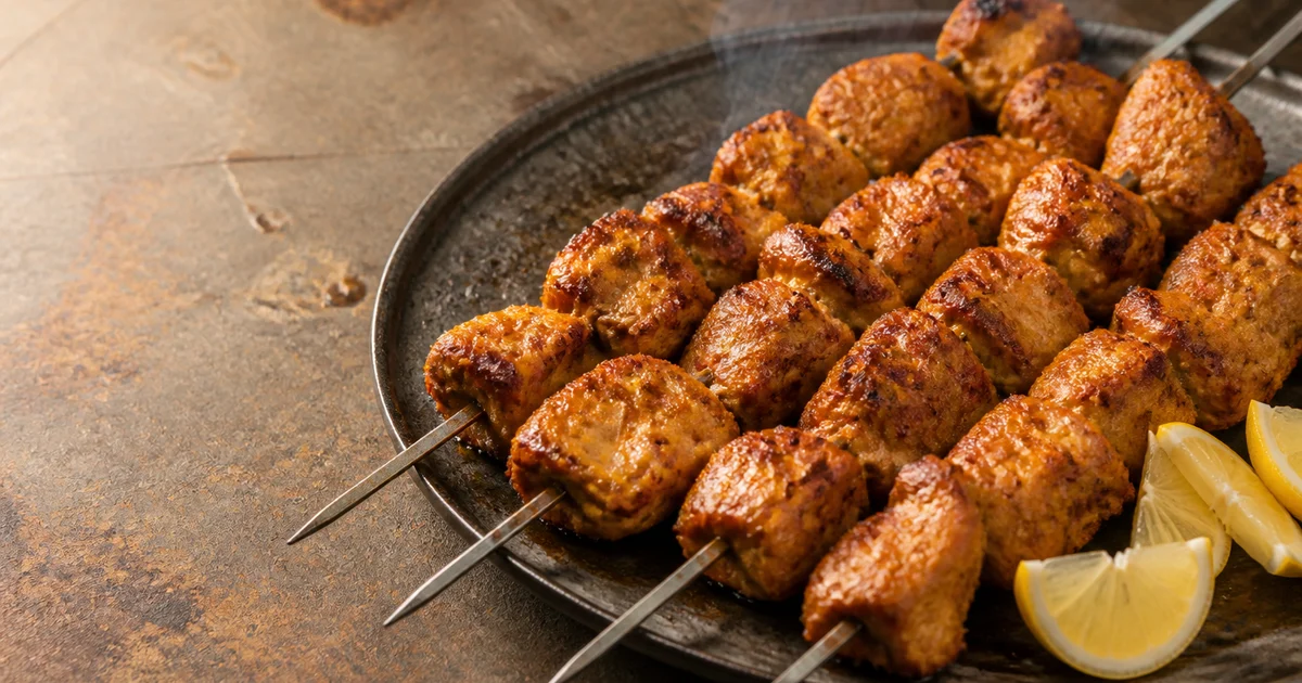 Char-grilled chicken boti kebab cubes with browned edges served on metal skewers beside onion, lemon and green chutney.