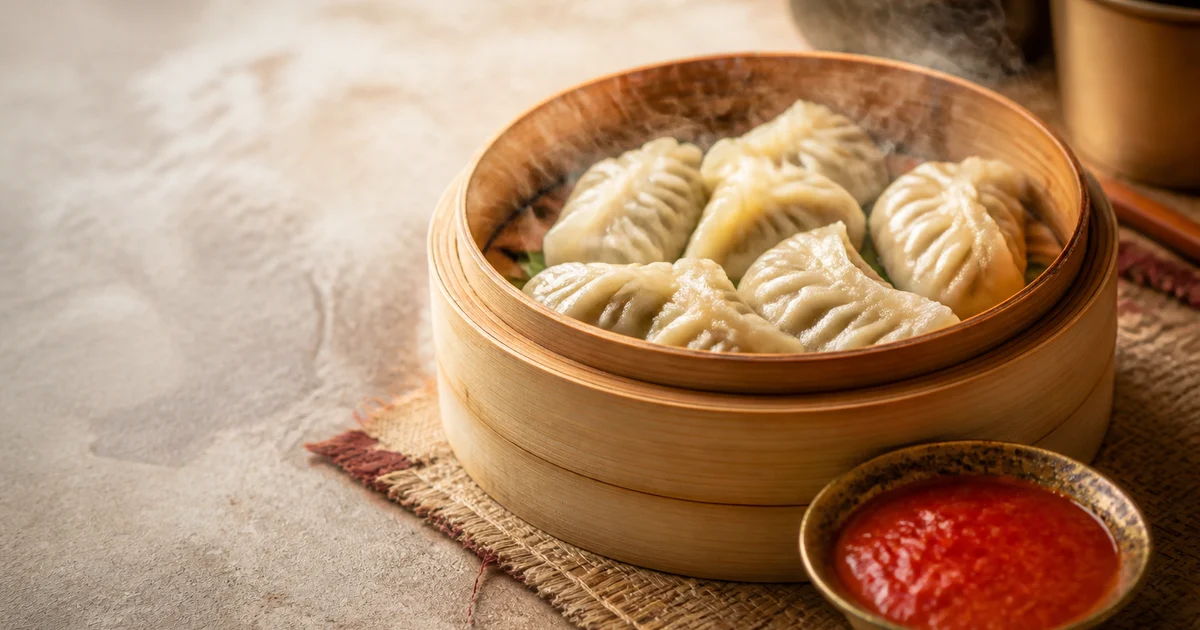 A plate of steamed chicken momos with pleated wrappers served beside red tomato chilli chutney.