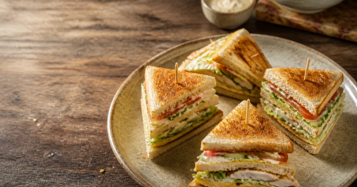 Four neatly cut chicken club sandwich triangles with toasted bread, egg, lettuce, cucumber and tomato secured with picks.