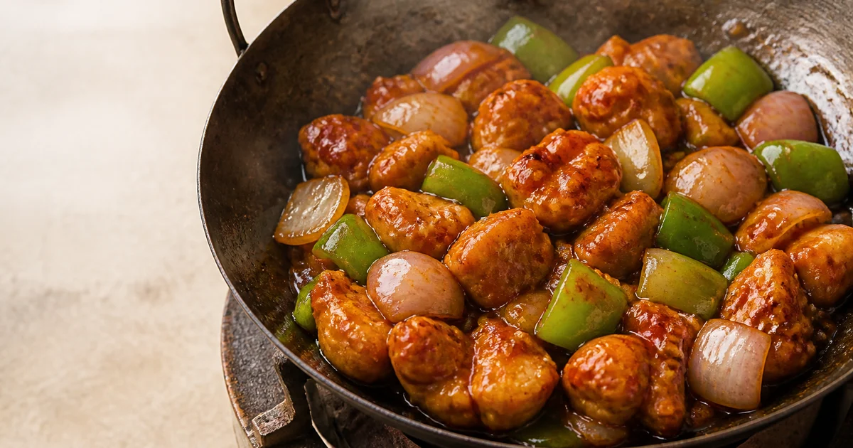 Crisp chilli chicken pieces tossed with glossy red sauce, onion petals, capsicum and green chillies in a serving bowl.
