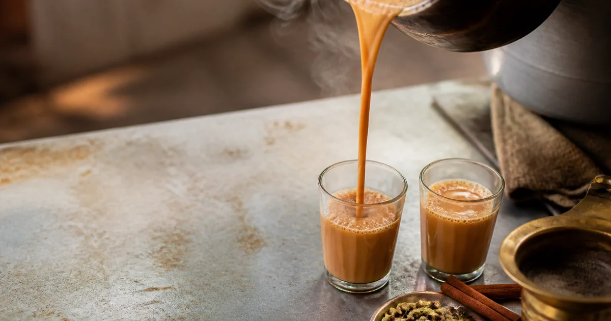 Fresh Bangladeshi masala cha being poured through a strainer into small serving cups beside whole spices and ginger.