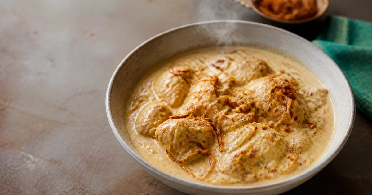 A serving bowl of pale Bangladeshi murgir korma with bone-in chicken pieces, creamy yoghurt gravy and green chillies beside polao.