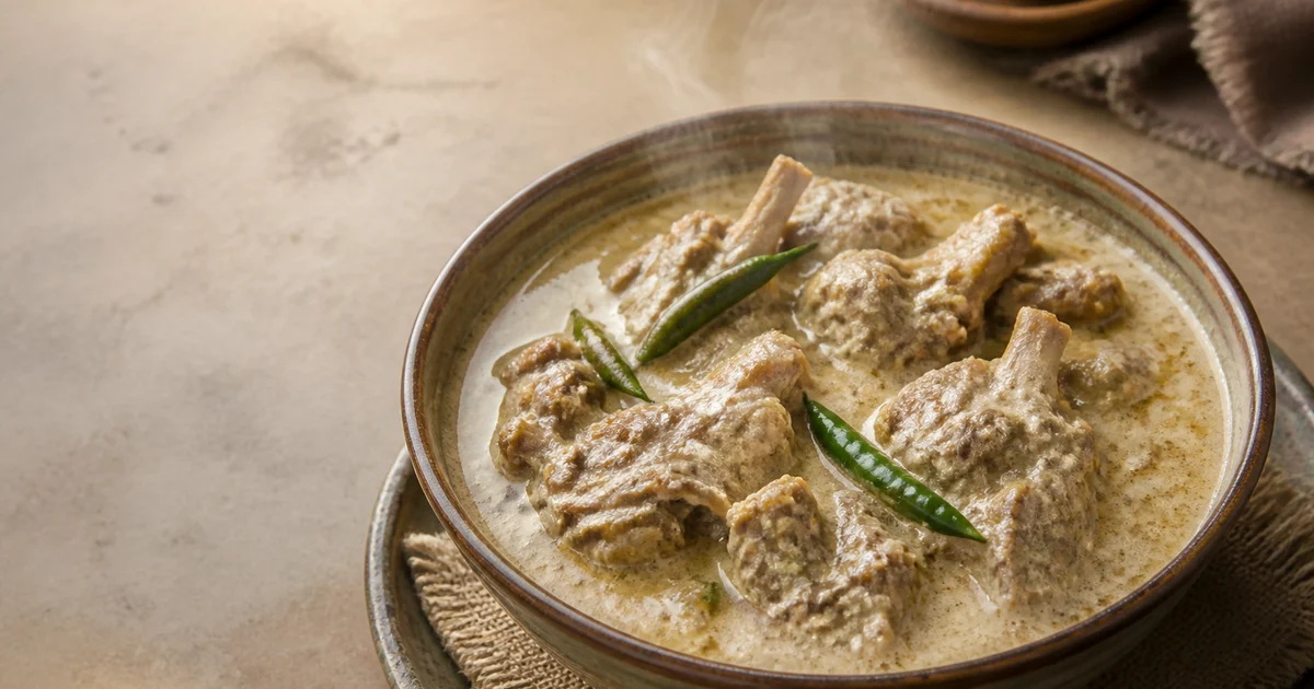A serving bowl of pale Bangladeshi mutton rezala with tender bone-in meat, creamy white gravy, whole spices and green chillies.