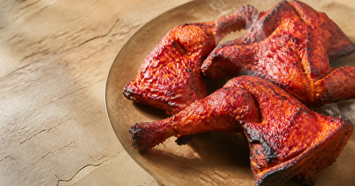 Charred red tandoori chicken leg quarters arranged on a metal serving tray with lemon wedges and sliced onions.